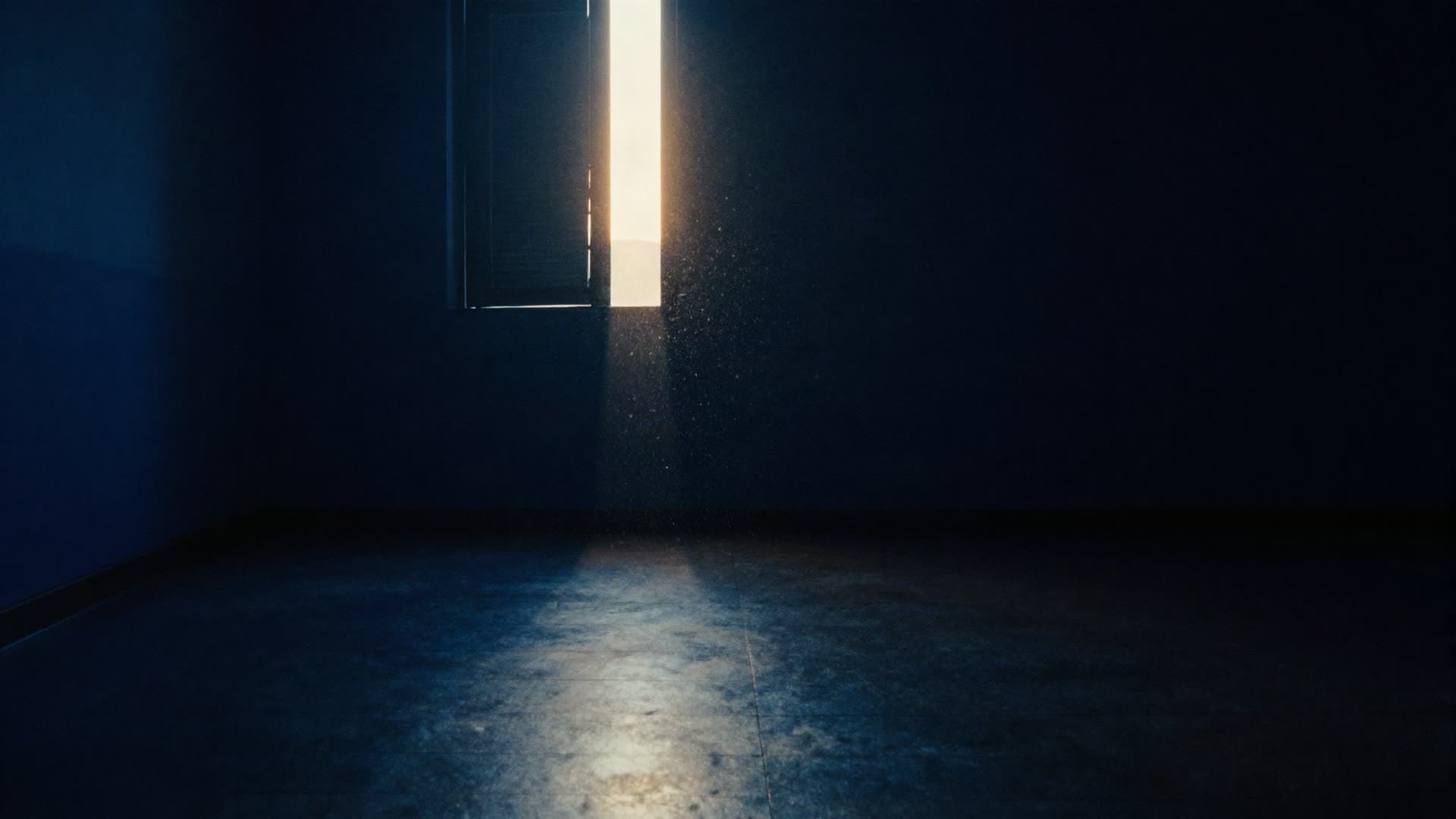A dark empty room with one narrow line of warm light entering through a gap, dust suspended in the beam.
