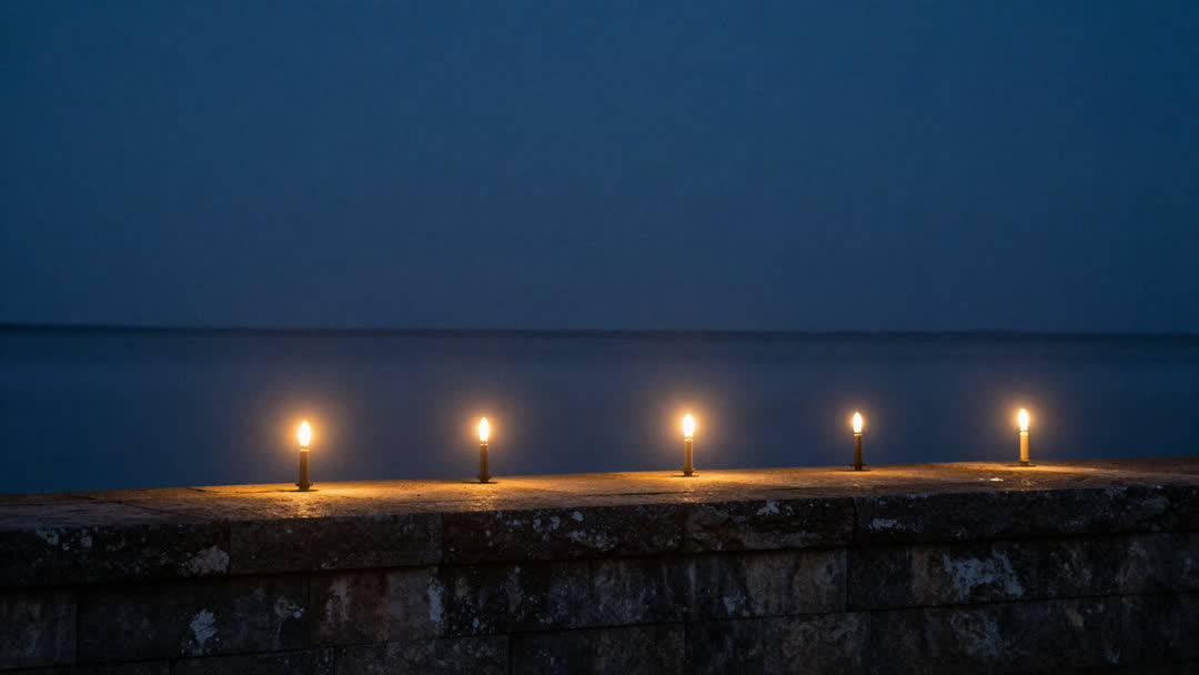 Seven small flames burning in a row along an ancient stone wall beside still water at first light.